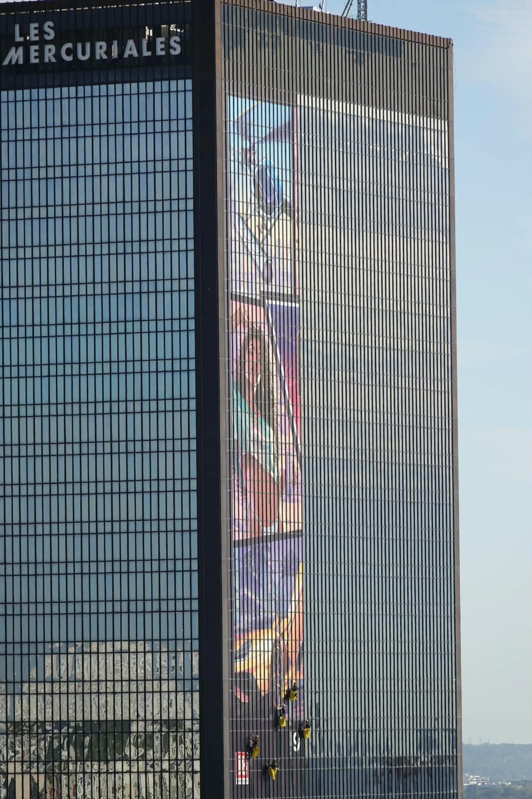 Façade d’une tour des Mercuriales avec une longue bande illustrée façon GTA VI et des cordistes suspendus qui la posent