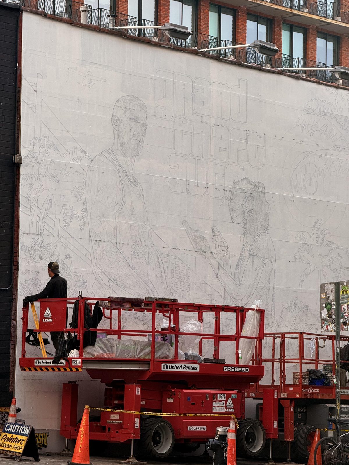Vue d’ensemble du mur peint à Williamsburg : esquisse de Jason et Lucia sous le logo Grand Theft Auto, nacelles élévatrices au pied du mur