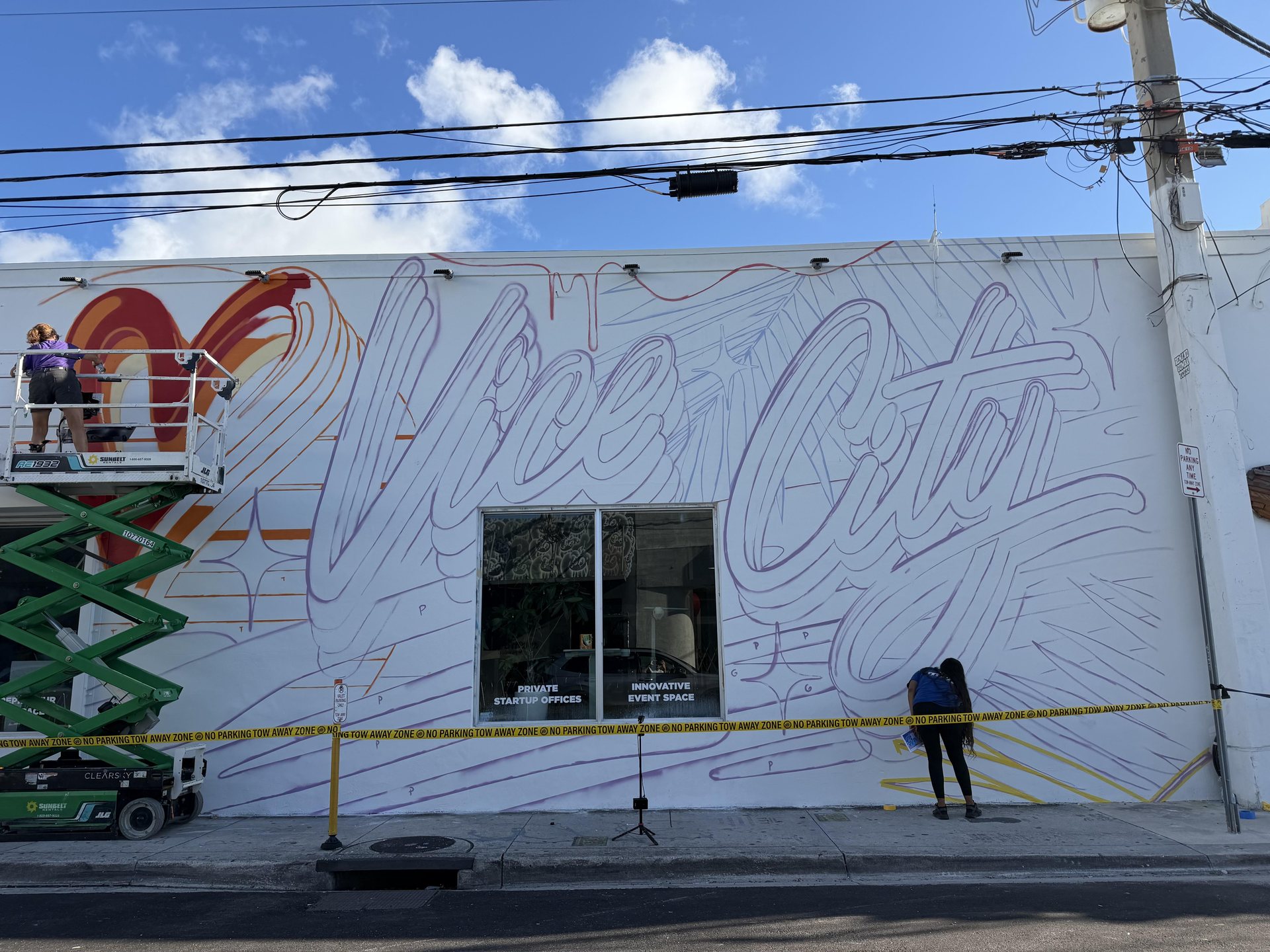 Fresque murale « Vice City » avec un cœur rouge en cours de peinture sur un mur blanc de Wynwood, à Miami, avec une nacelle et une peintre