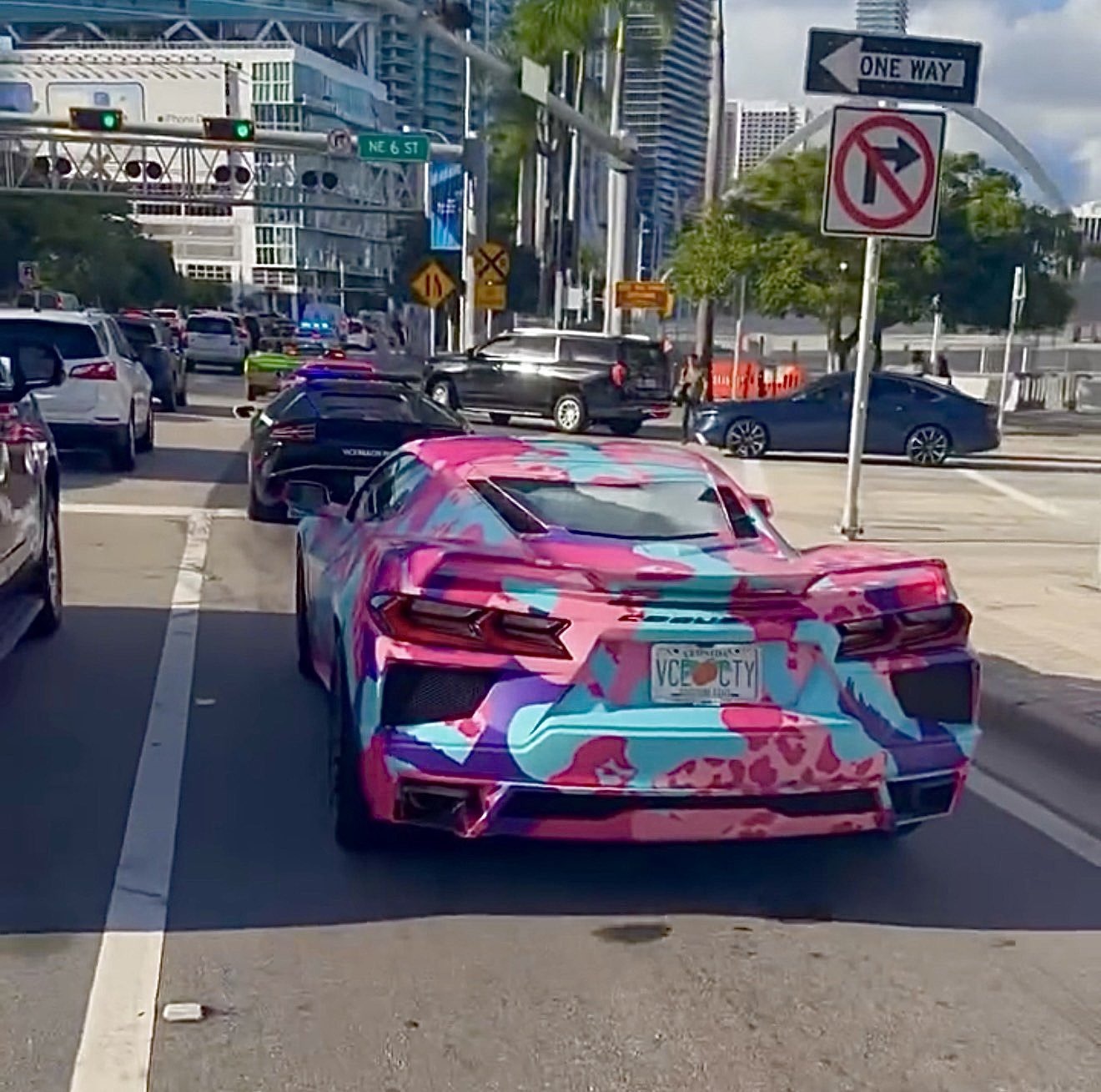 Une Corvette recouverte d’un camouflage rose, bleu et violet, plaque VCE CTY, dans une rue de Miami devant la Lamborghini de police
