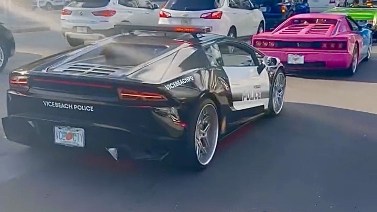 Une Lamborghini noire et blanche aux couleurs de la Vice Beach Police, plaque VCE CTY, roule dans le centre de Miami suivie d’une voiture rose