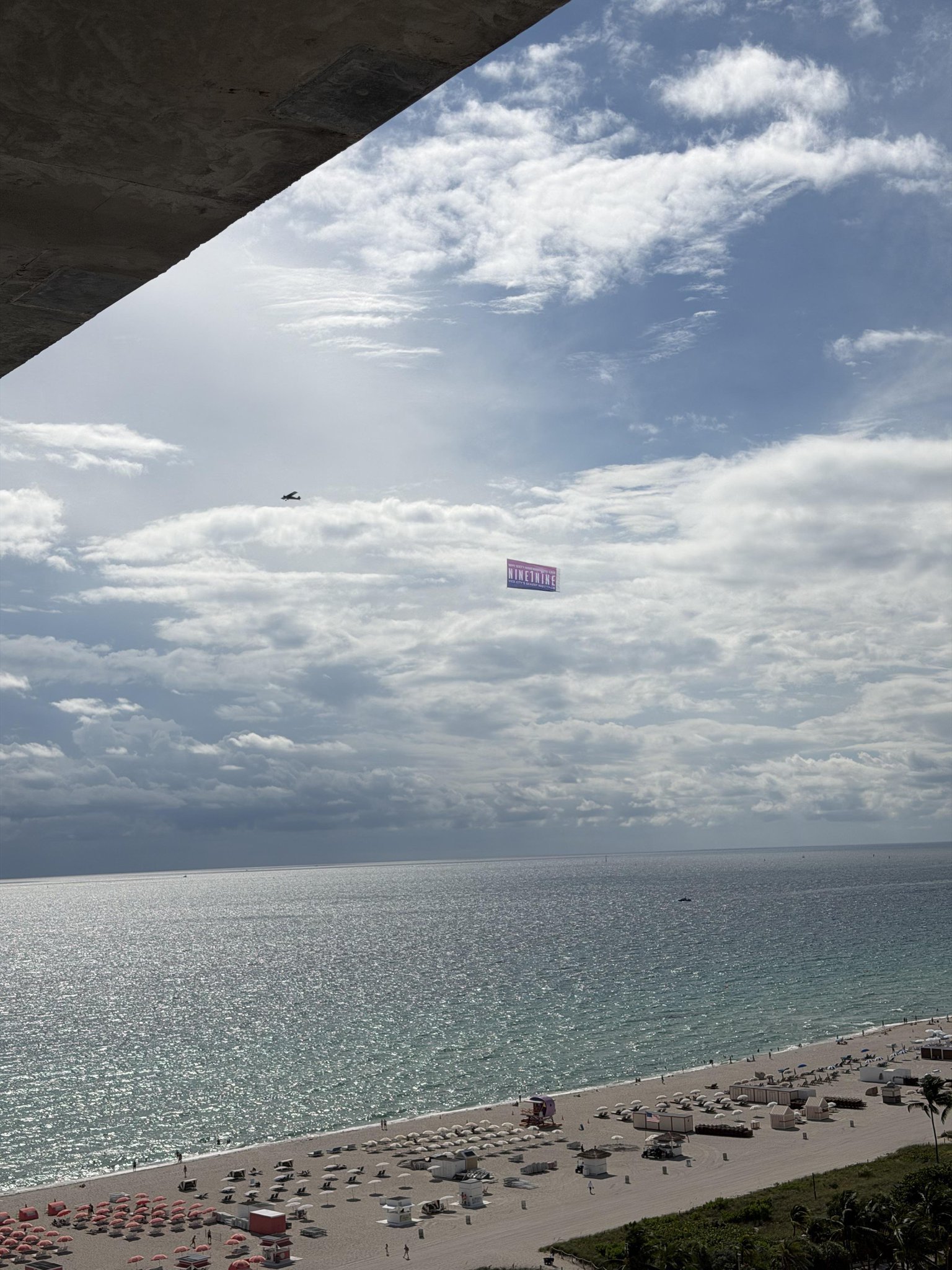 Vue en hauteur de South Beach : un petit avion tracte la banderole NINE1NINE au-dessus de l’océan, la plage et ses parasols en contrebas