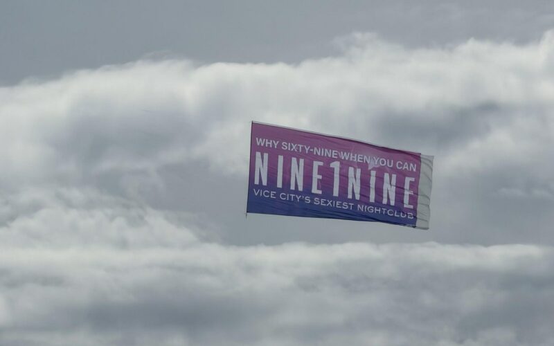 Banderole rose et violette « Why sixty-nine when you can NINE1NINE, Vice City’s sexiest nightclub » tractée dans le ciel nuageux de Miami Beach