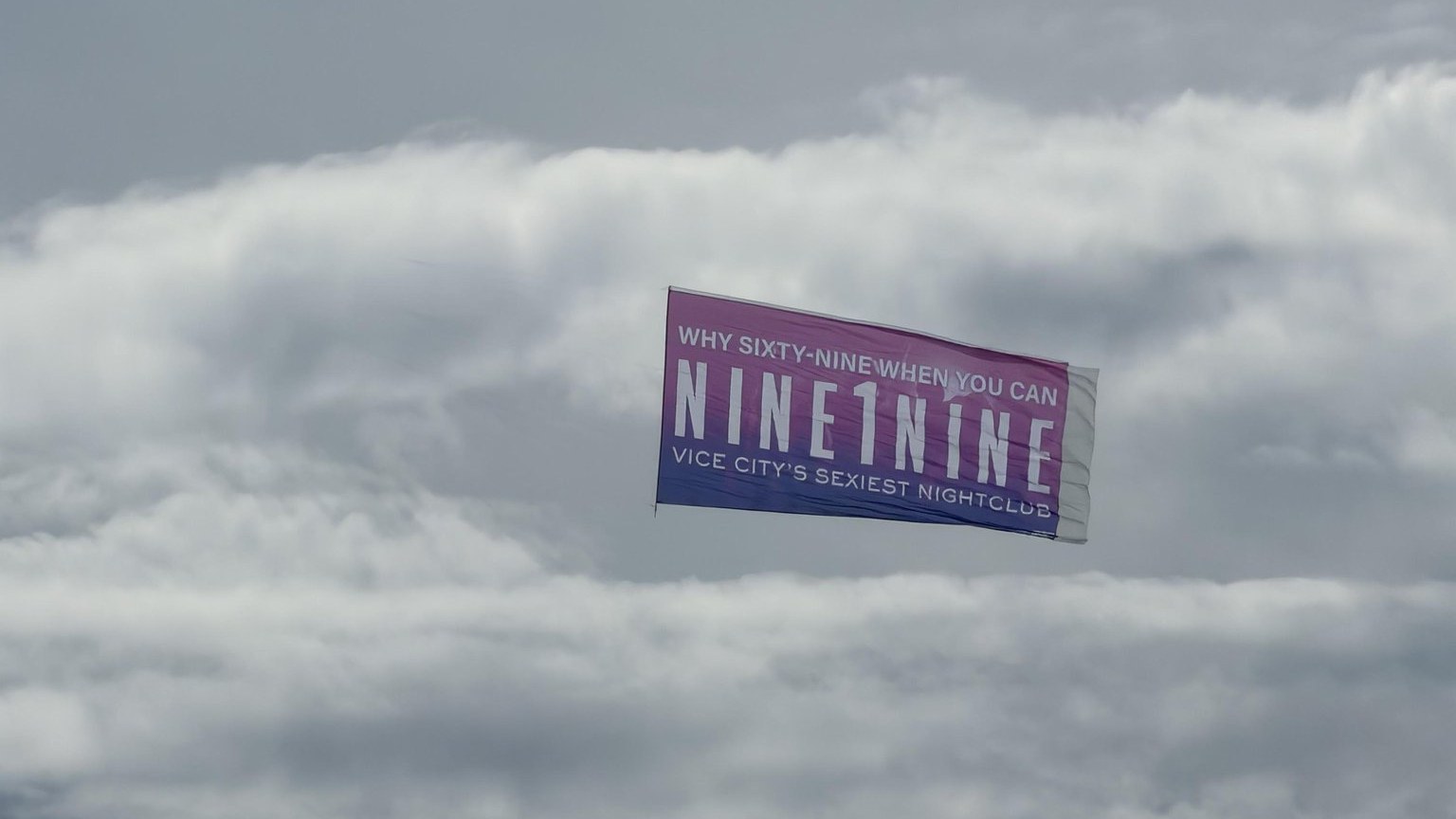 Banderole rose et violette « Why sixty-nine when you can NINE1NINE, Vice City’s sexiest nightclub » tractée dans le ciel nuageux de Miami Beach