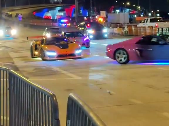 De nuit, une supercar orange et une voiture rose aux néons bleus roulent sur une route bouclée près du port de Miami, gyrophares de police en arrière-plan
